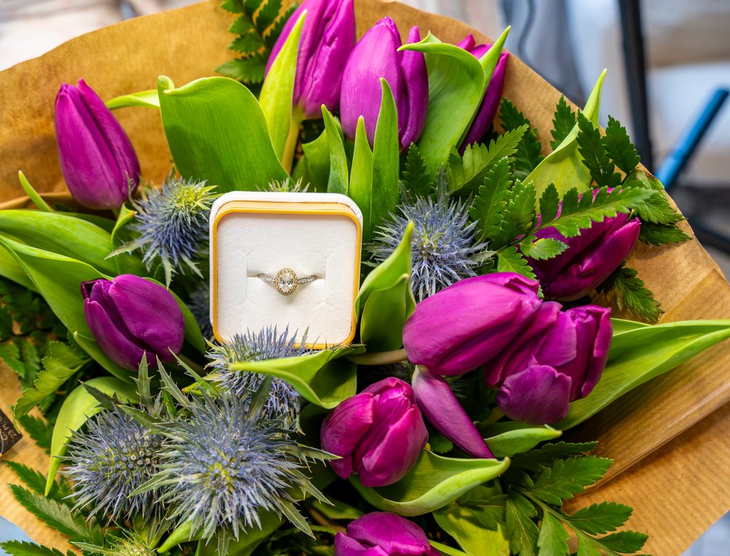 Une bague de fiançailles dans son écrin, posée au cœur d’un bouquet de tulipes magenta et de chardons bleus.