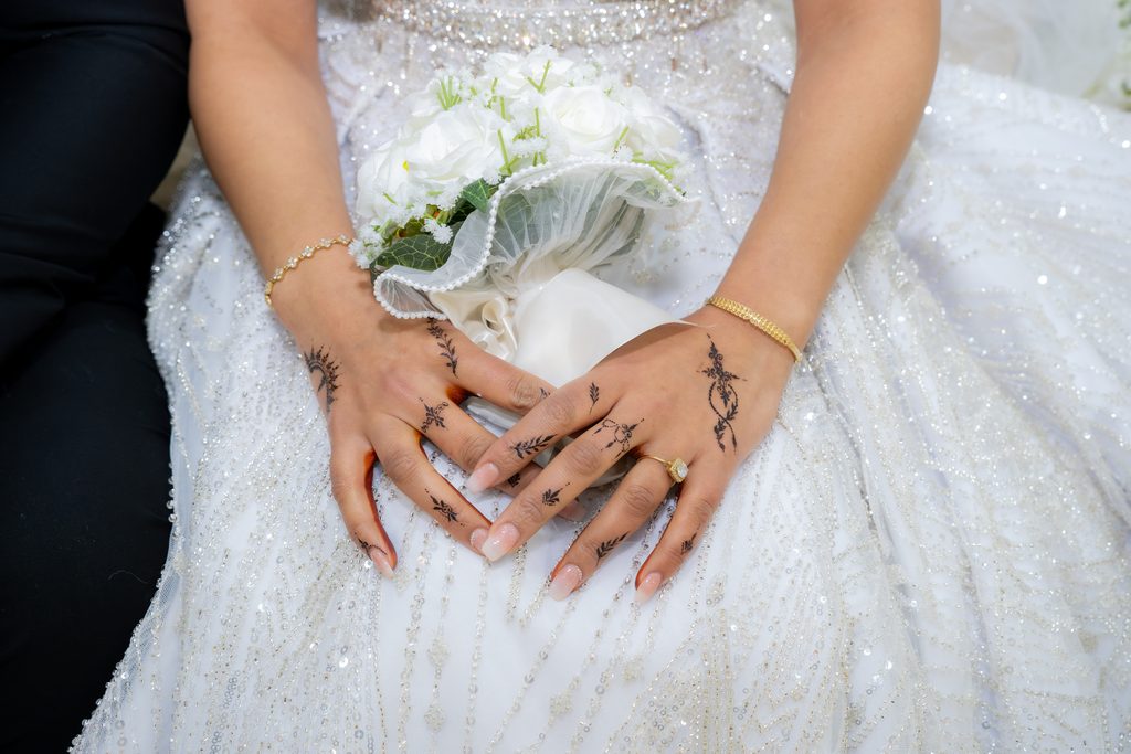 Les mains de la mariée ornées de henné, posées sur sa robe brodée, autour d’un bouquet blanc.