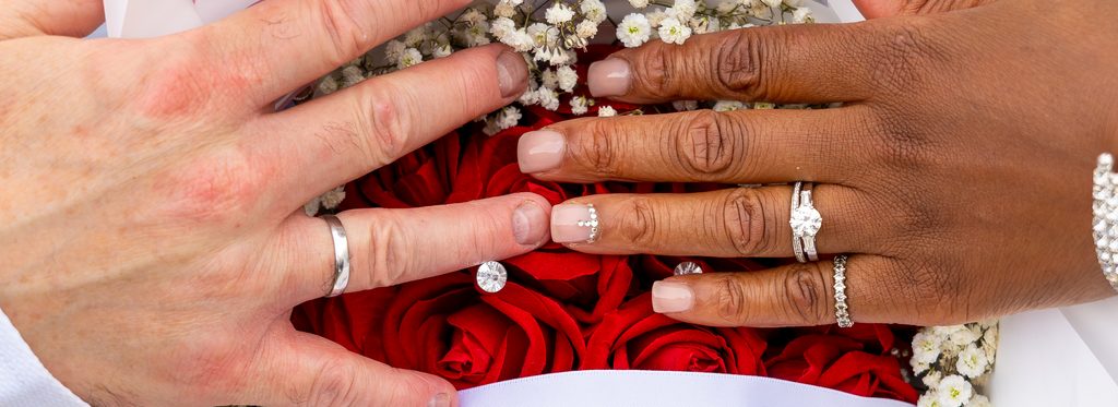 Deux mains posées côte à côte sur un bouquet de roses rouges et de gypsophile, alliances visibles.